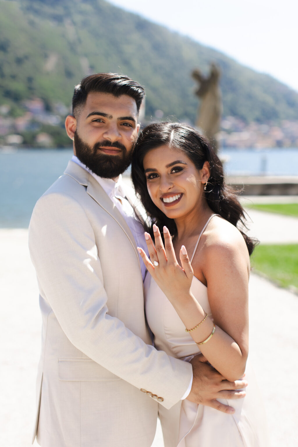 A newly engaged couple embracing and smiling at the camera while the bride-to-be shows her diamond ring, on a lakeside lawn on Lake Como.
