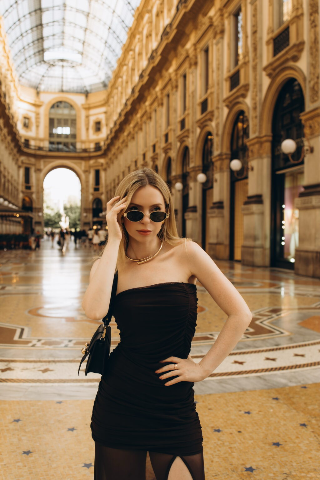 Woman in a strapless dark dress, gold collar necklace, and oval sunglasses posing beneath the glass vault of Galleria Vittorio Emanuele II in Milan, on the star-patterned mosaic floor
