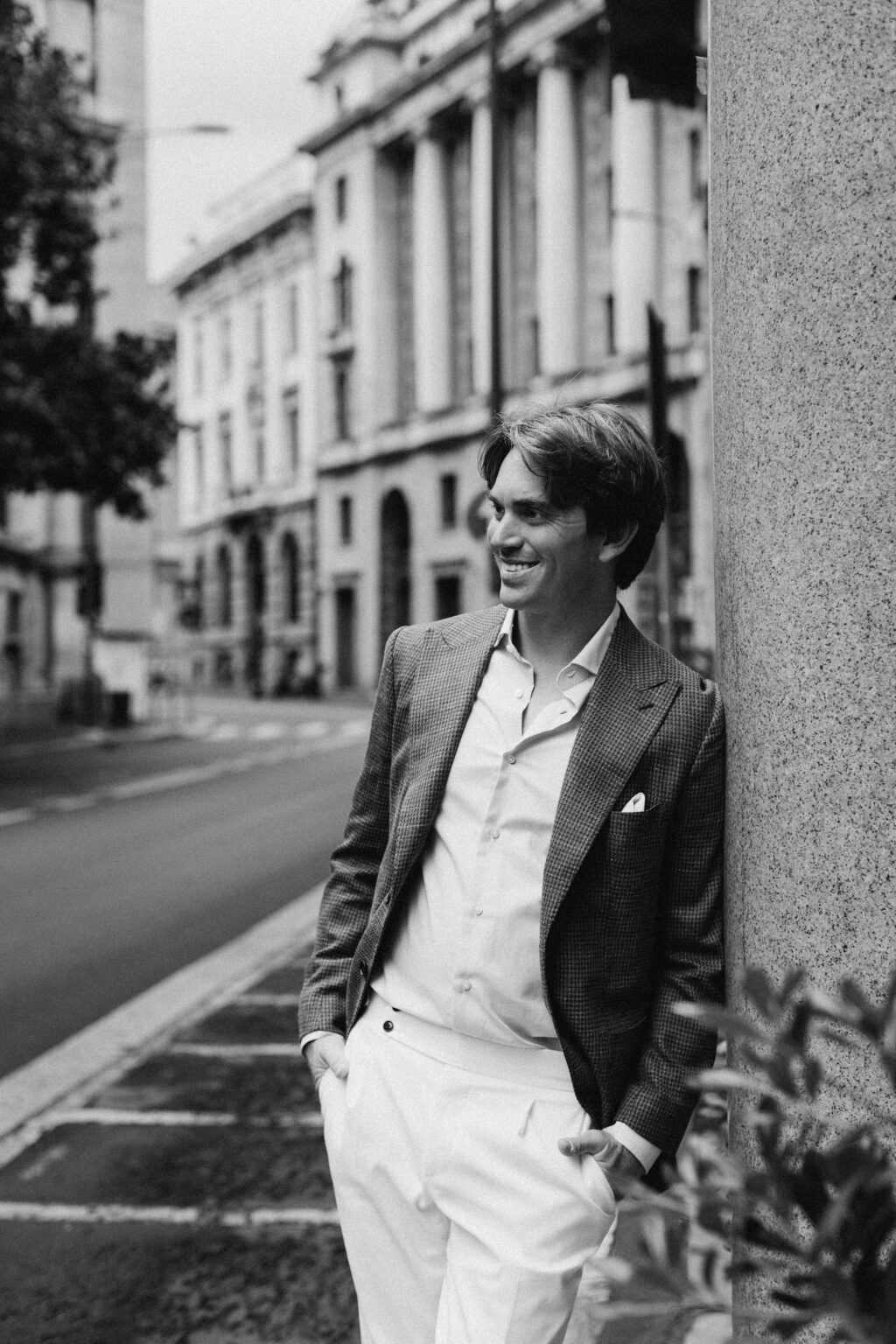 A black and white photo of a smiling man leaning against a stone column on a Milan street, a neoclassical colonnade behind him.