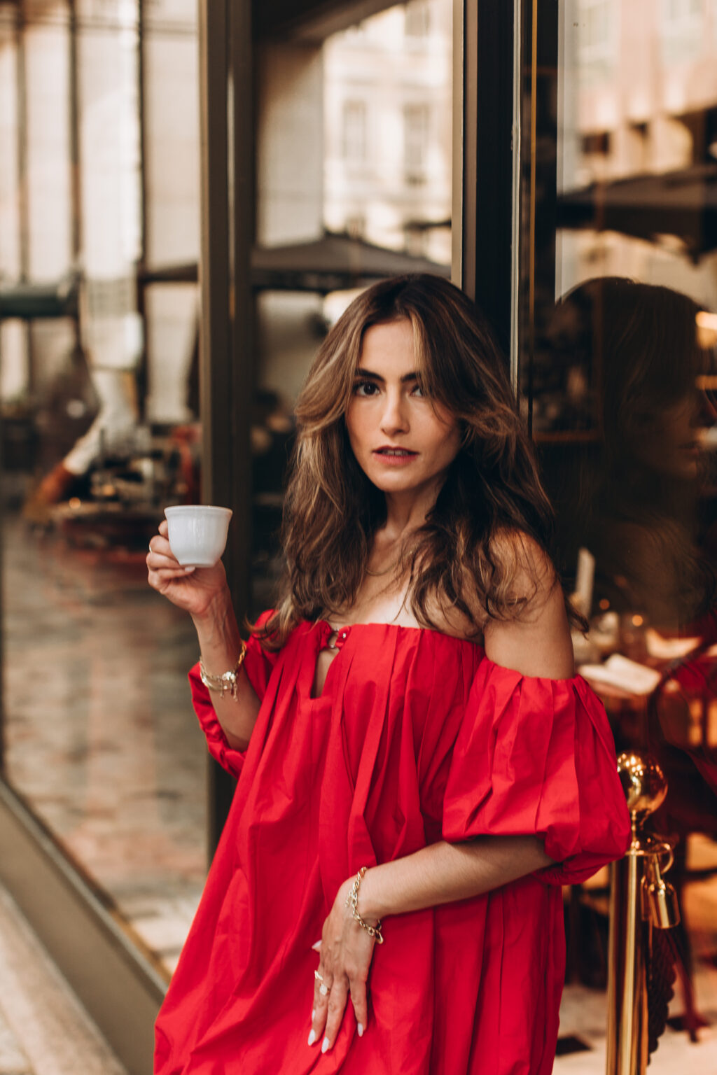 Woman in a red off-shoulder dress holds an espresso cup beside a café window in Milan.