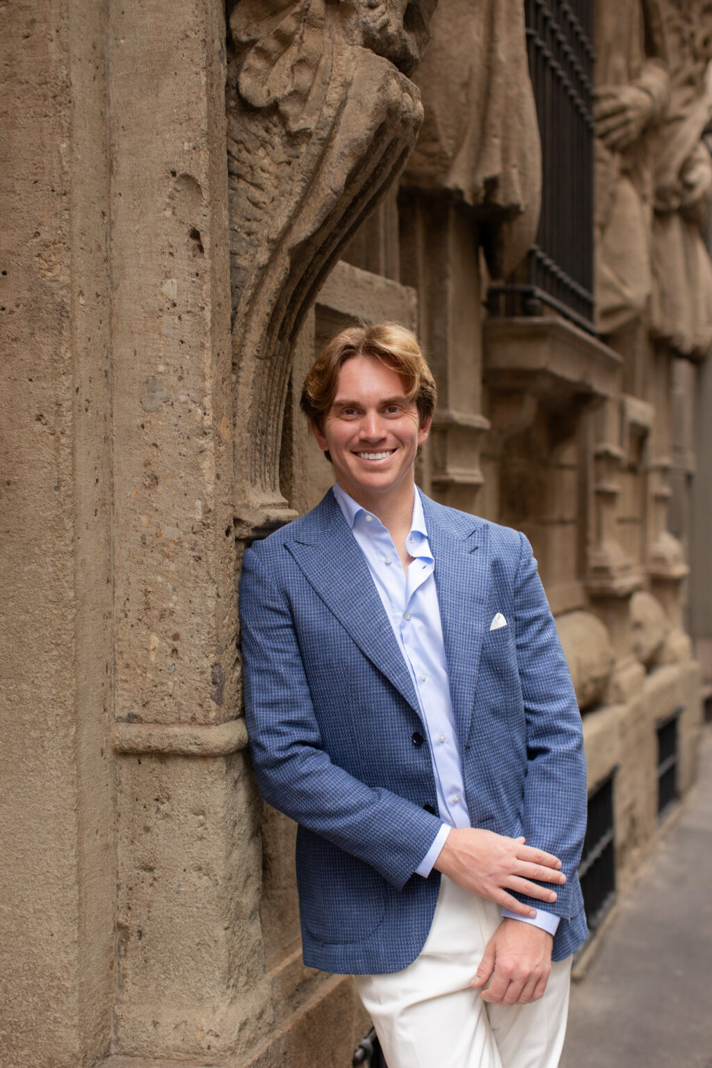 A smiling man in a blue jacket leaning against the sculpted stone facade of Casa degli Omenoni in central Milan.