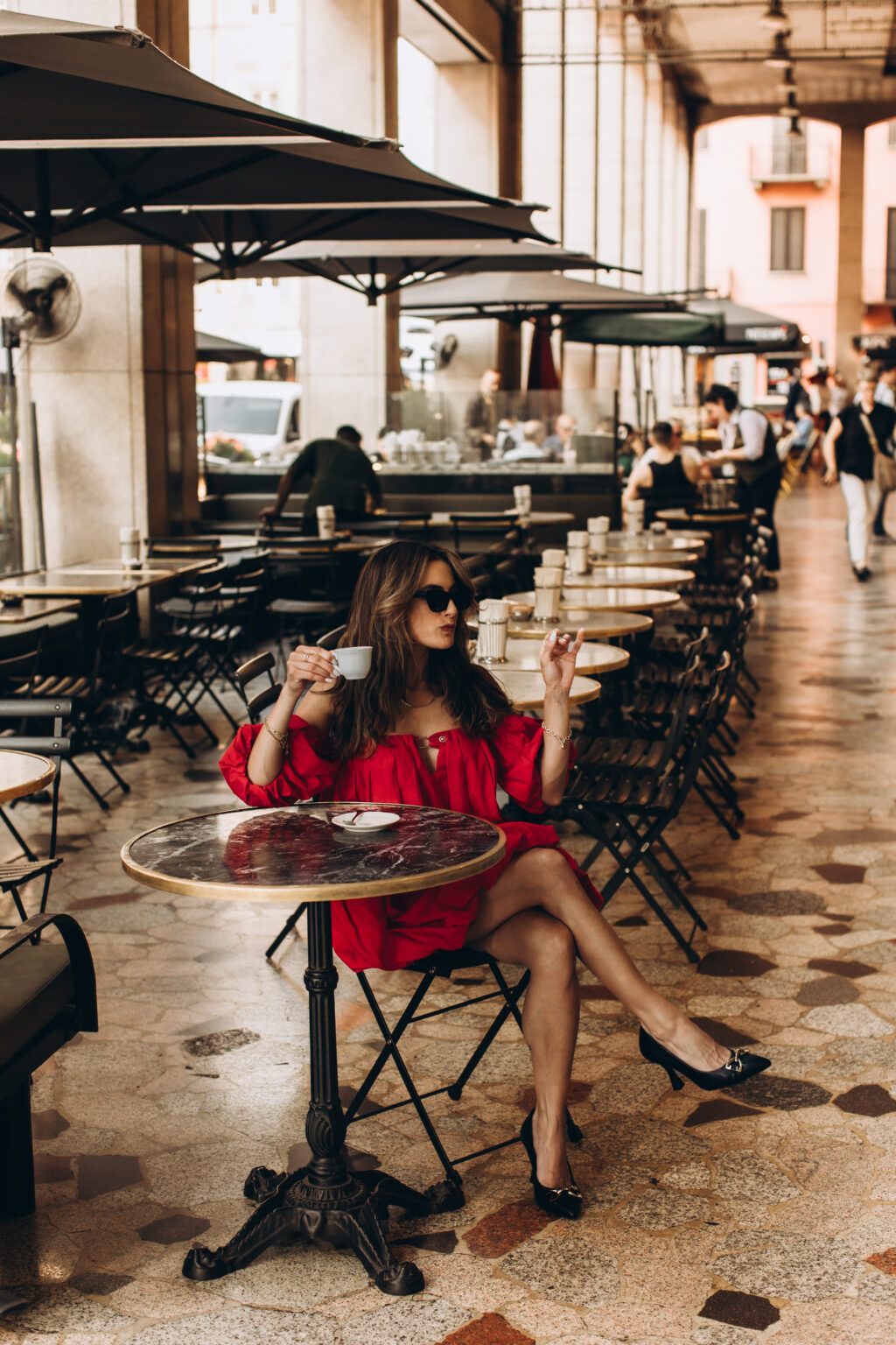 Woman in a red off-shoulder dress sips espresso at a marble café table under a Milan portico.