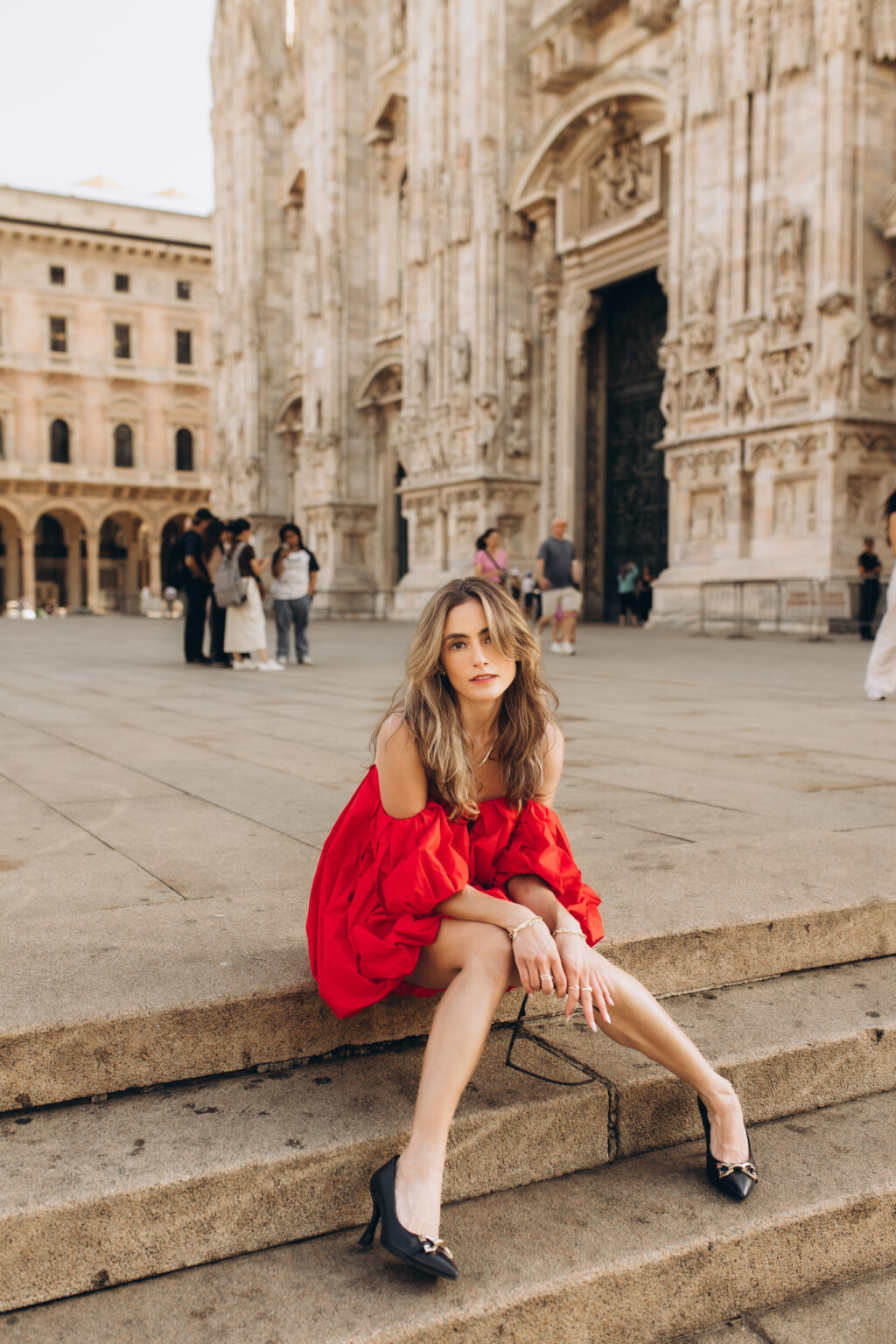 Woman in a red off-shoulder dress sits on the steps of the Duomo di Milano in Piazza del Duomo in Milan.