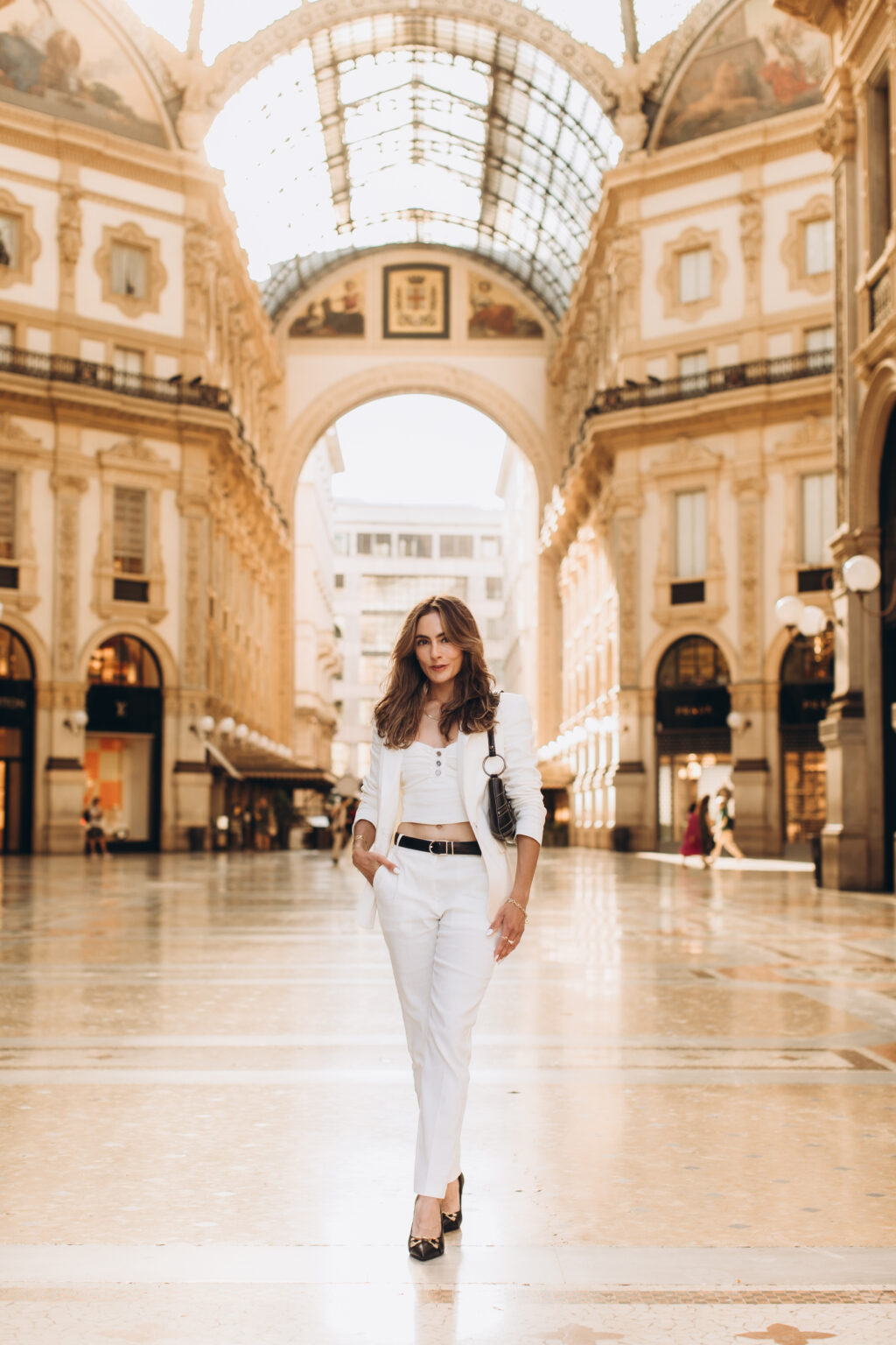 Woman in a white suit stands beneath the golden arch of the Galleria Vittorio Emanuele II during a photoshoot for Instagram in Milan.