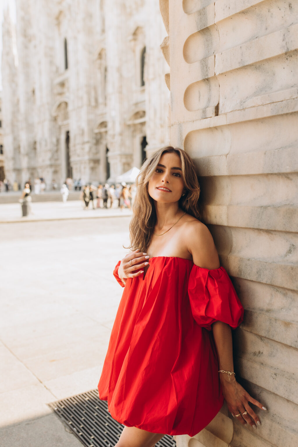Woman in a red off-shoulder dress leans against a marble wall with the Duomo di Milano behind her in Milan.