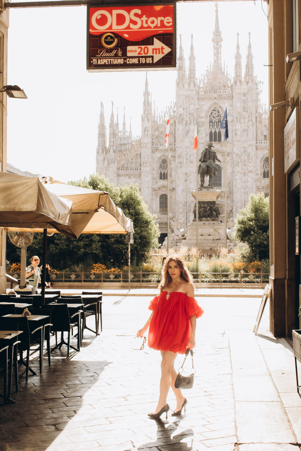 Woman in a red dress walks under a portico with the Duomo di Milano rising in the background in Milan.