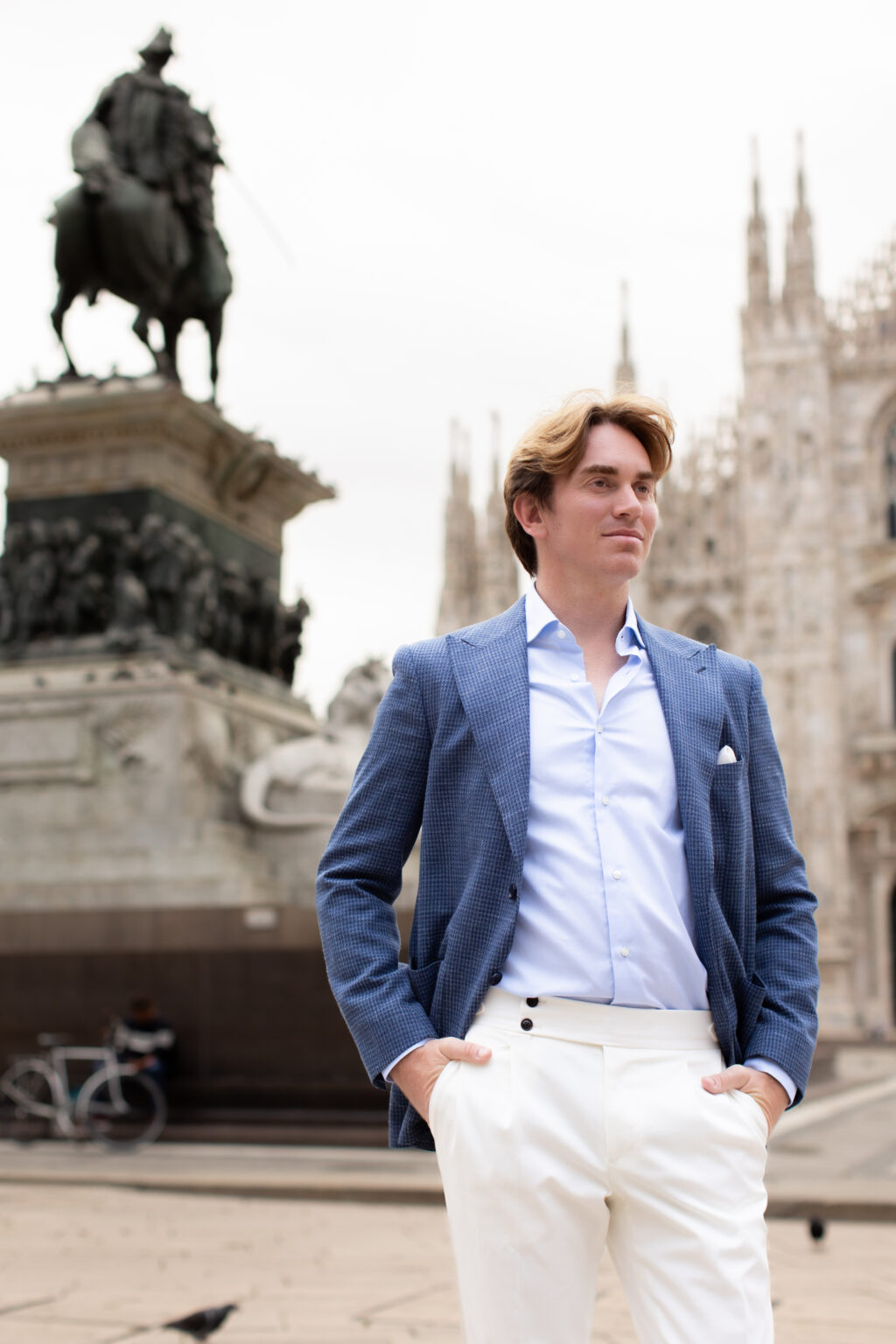 A man in a blue blazer standing before the equestrian Monument to Victor Emmanuel II, with the Duomo di Milano to the side in Milan.