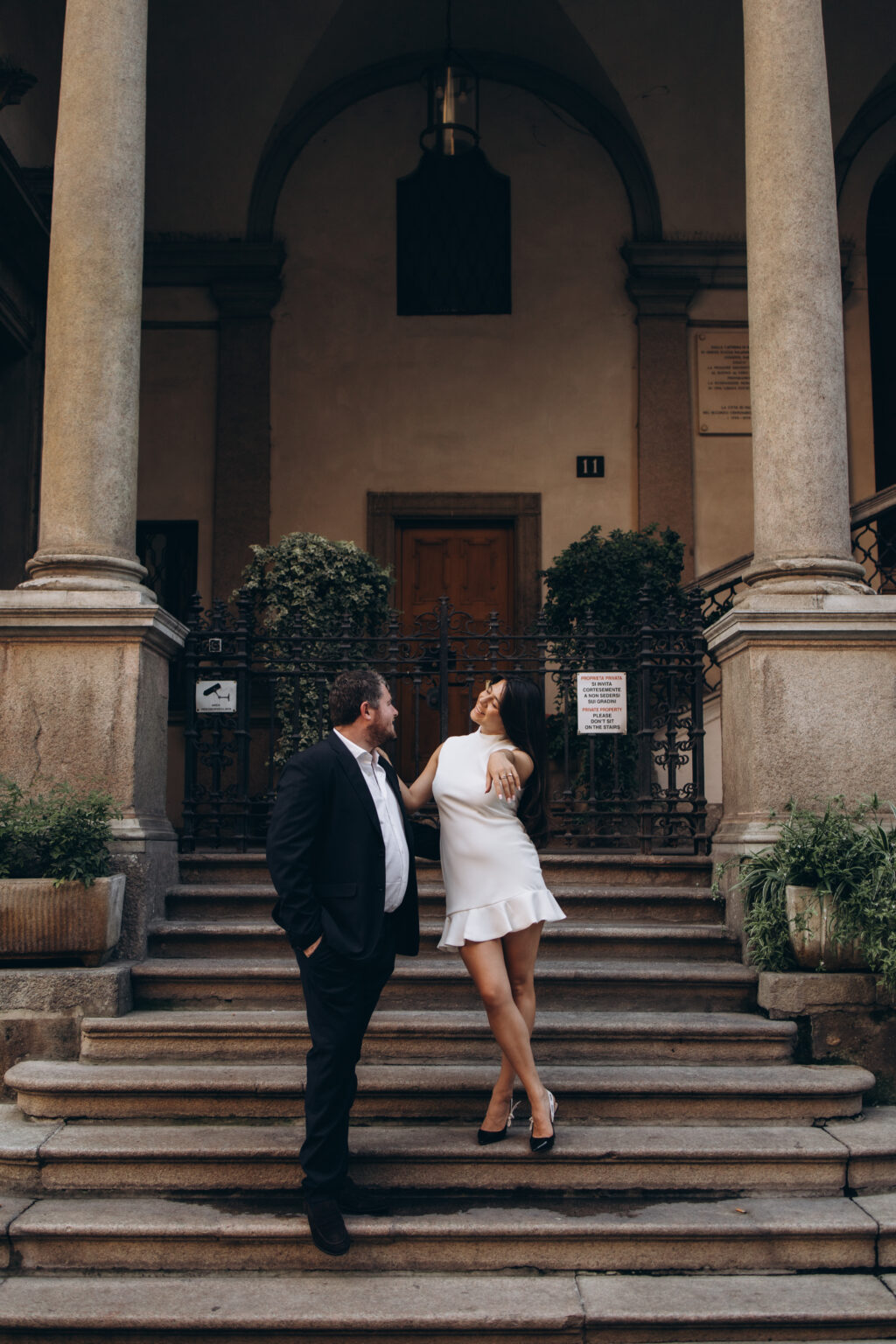 Woman in a white dress playfully leaning toward her fiance while both laugh on the steps of a columned courtyard in Milan