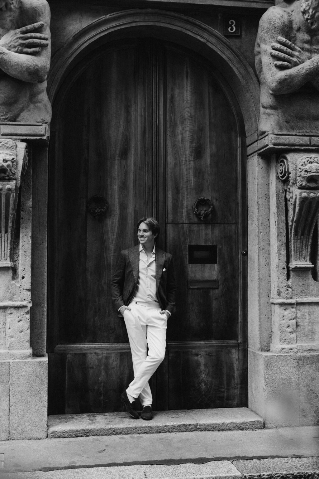 A black and white photo of a man leaning against the grand carved wooden door of Casa degli Omenoni, framed by telamon sculptures, in Milan.