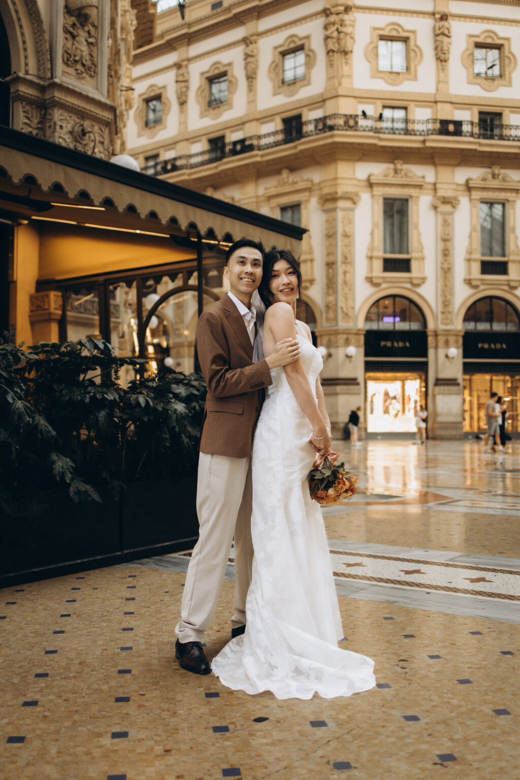 Groom-to-be embracing his smiling bride-to-be from the side as both look at the camera in the Galleria Vittorio Emanuele II in Milan