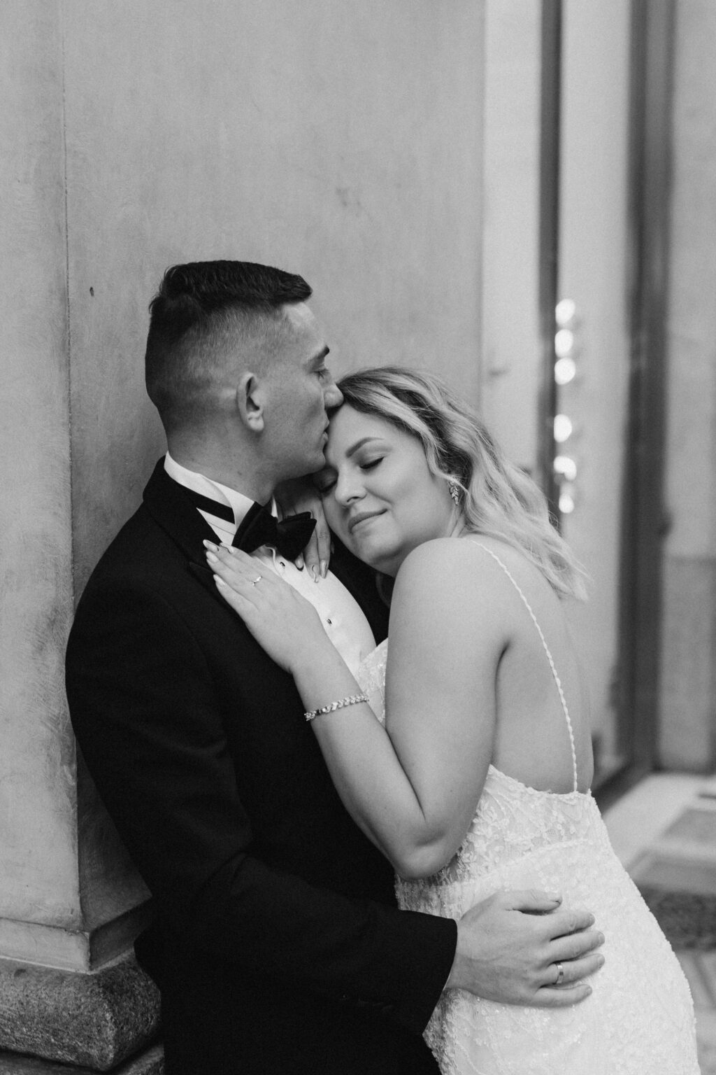 A black and white portrait of him kissing her forehead as they lean against a column, her hand resting on his chest.