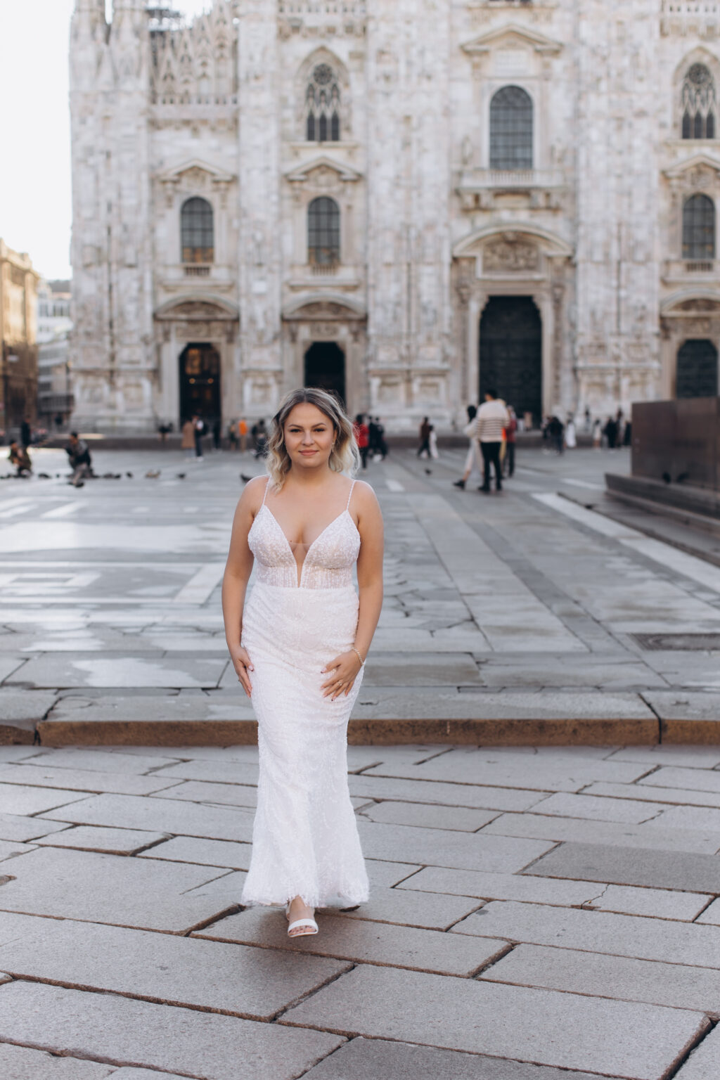 She walks toward the camera in a fitted beaded gown on Piazza del Duomo, the marble facade of the Milan Cathedral behind her.