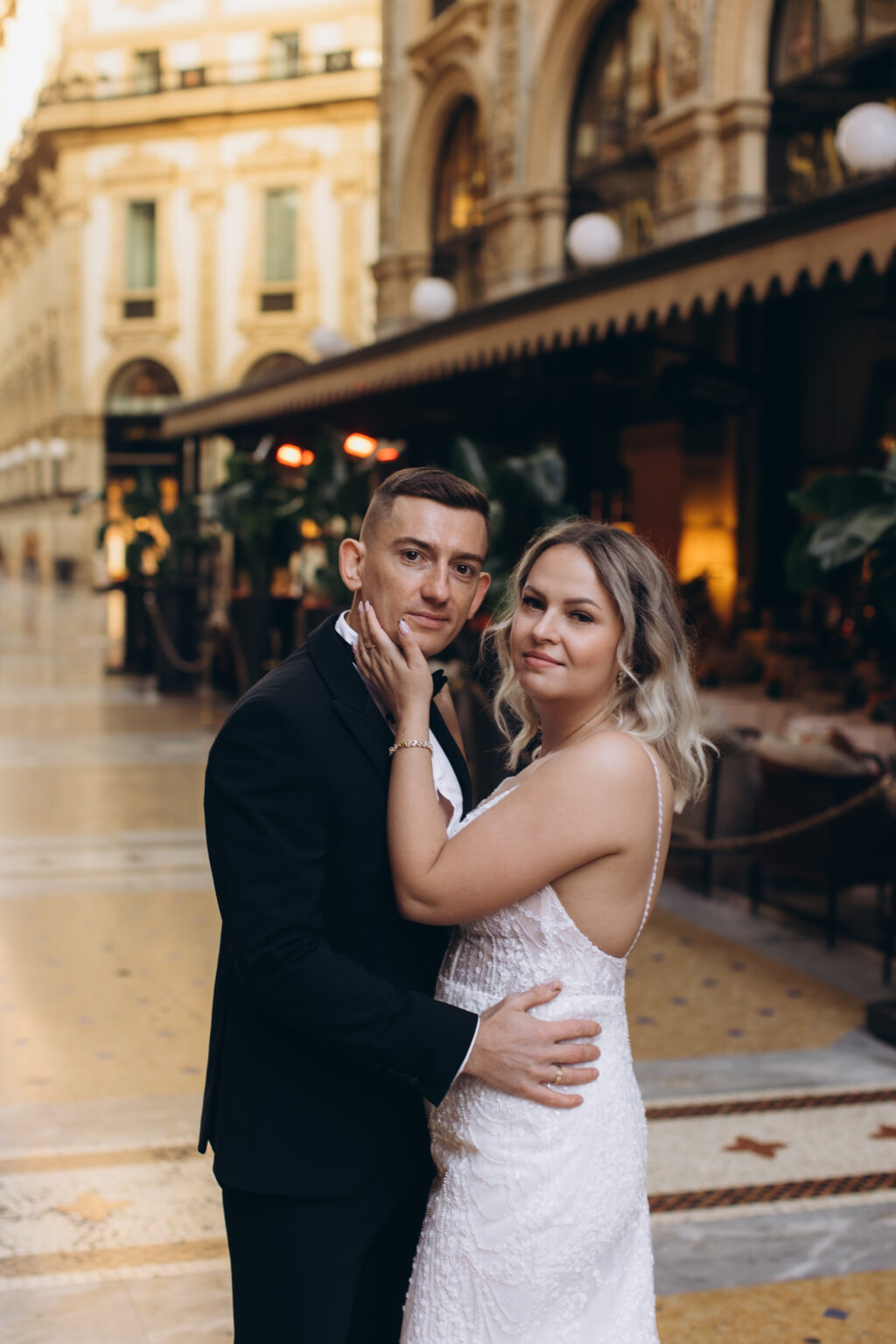 She touches his chin in a close embrace inside the Galleria Vittorio Emanuele II, warm restaurant lights and arcades behind them.