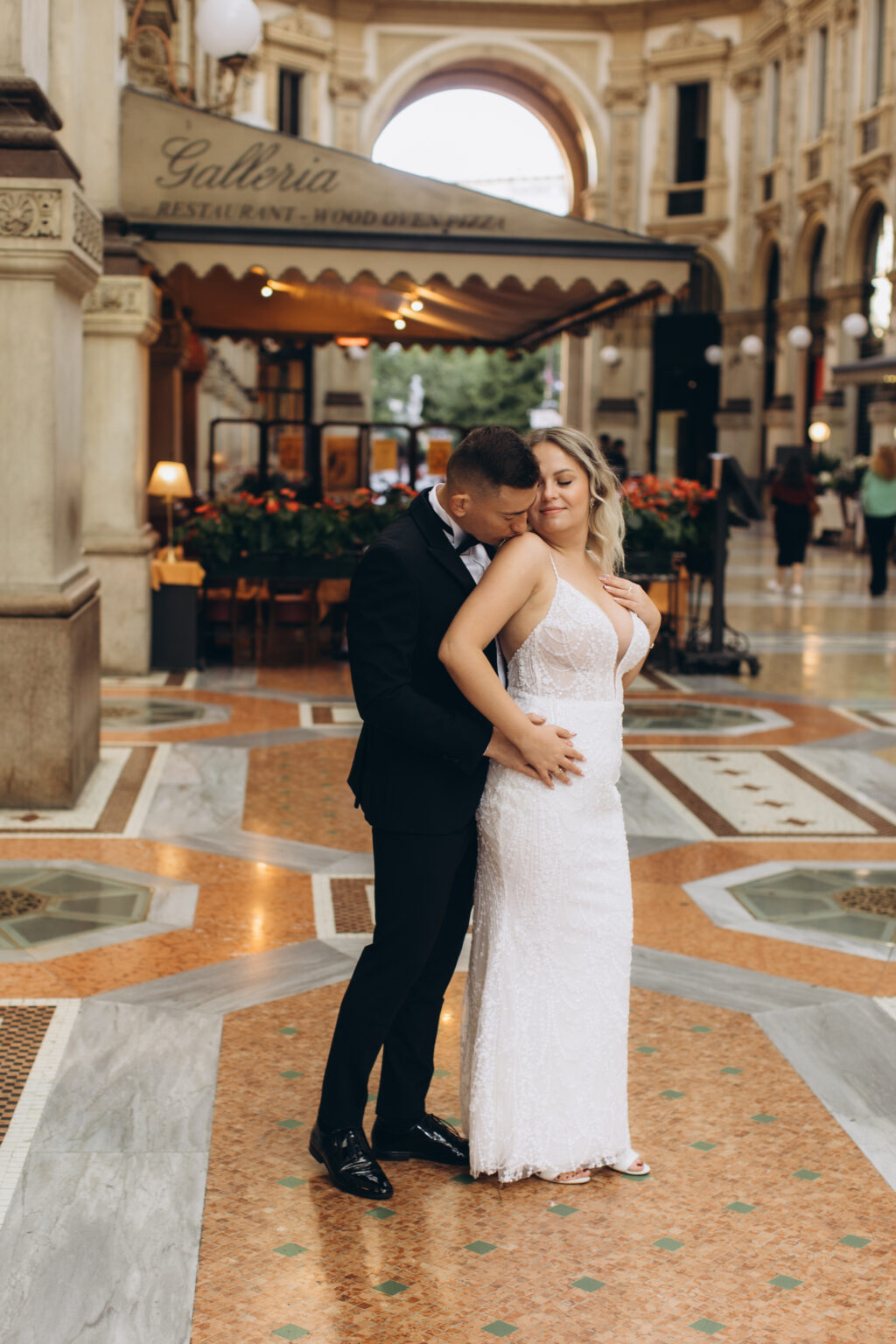 He kisses her shoulder in a close embrace on the mosaic floor of the Galleria Vittorio Emanuele II, its arch and café awnings behind them.