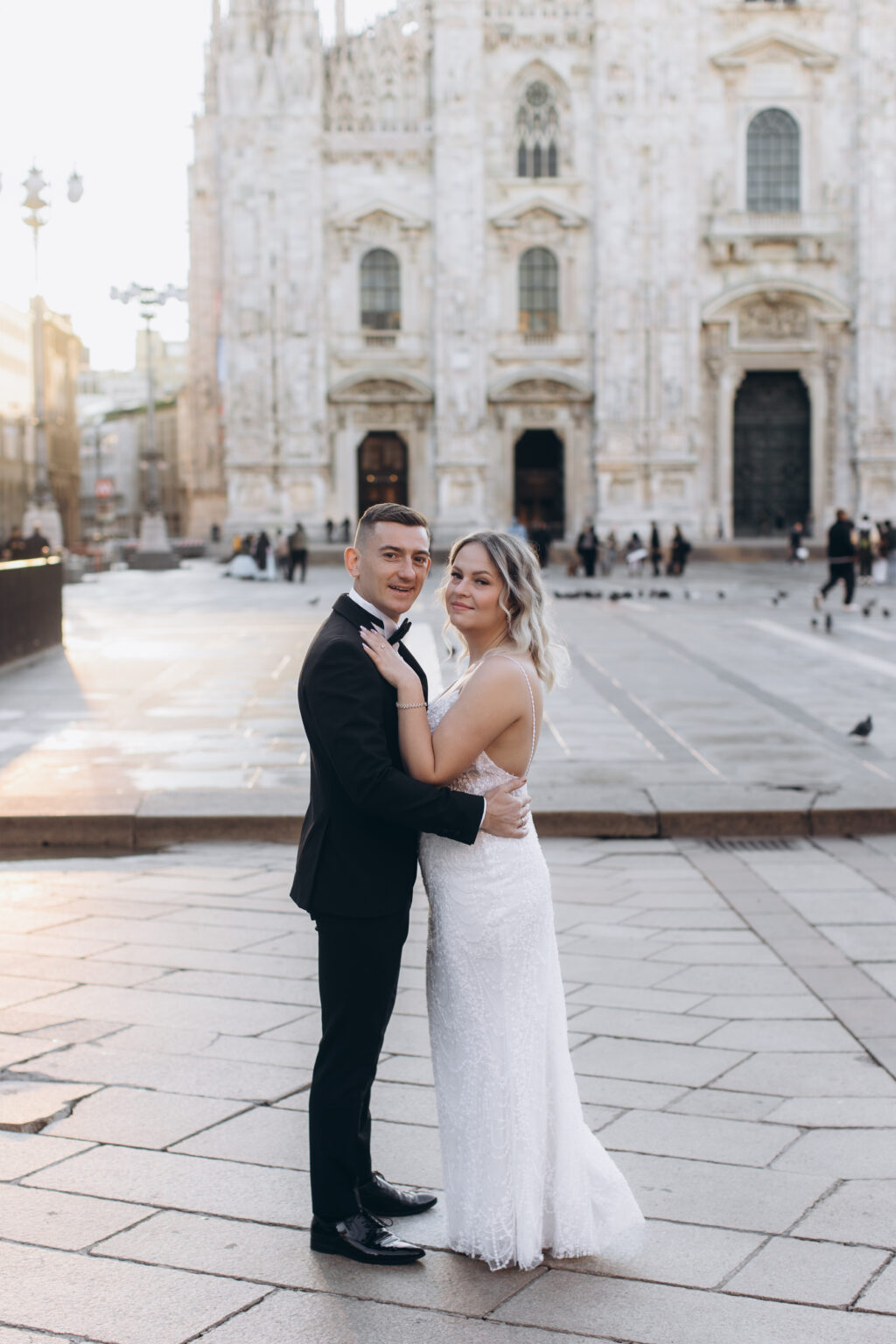She in a beaded gown and he in a black tuxedo embrace on Piazza del Duomo at golden hour, the marble facade of the Milan Cathedral behind them.