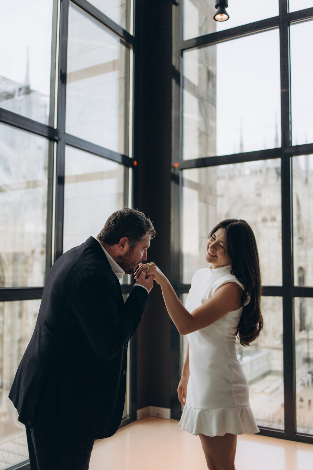Man kissing the hand of his fiancee, who wears her new engagement ring, by the corner windows of the Museo del Novecento in Milan