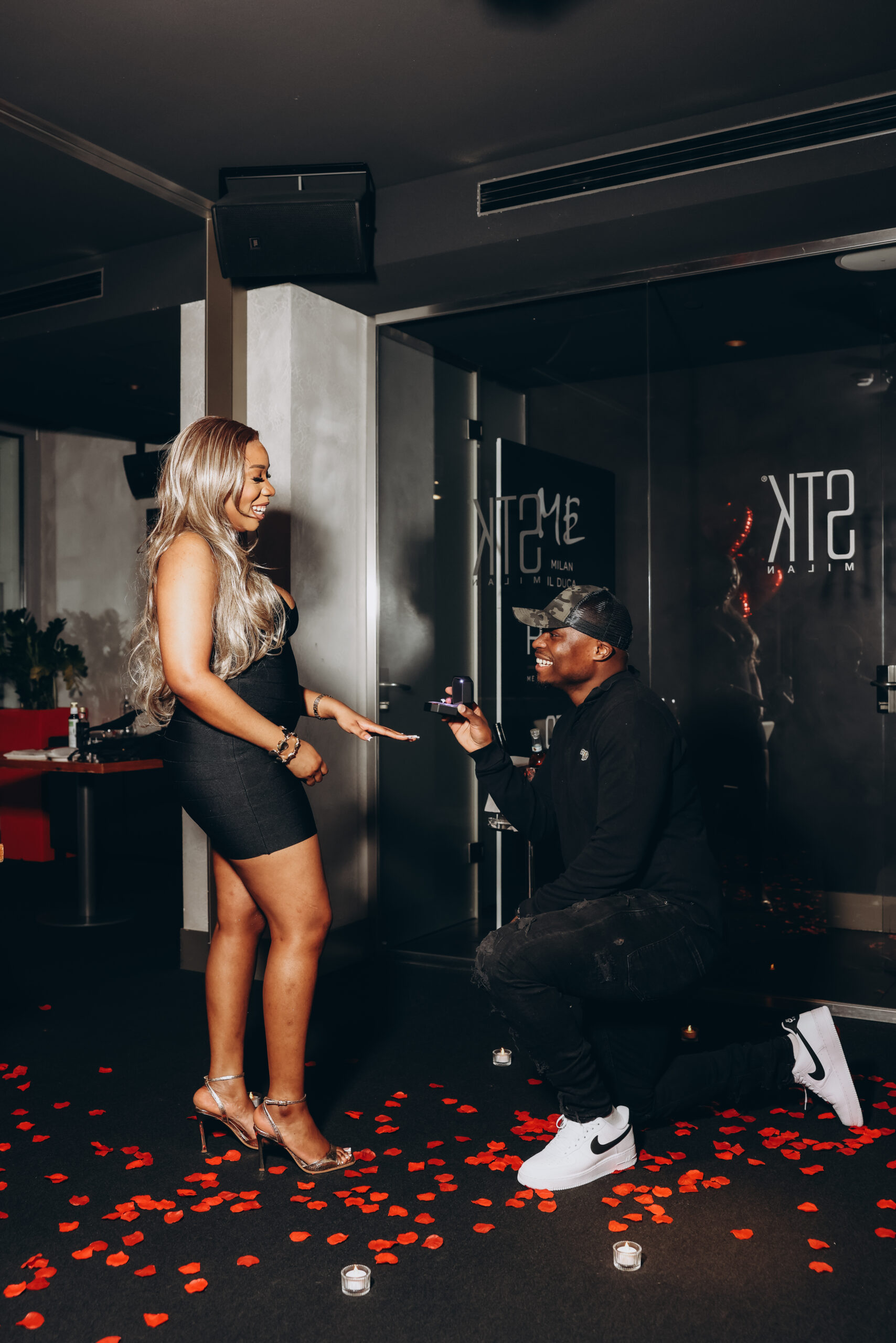 A man kneels and presents an engagement ring to his smiling partner among rose petals and candles at a Milan restaurant
