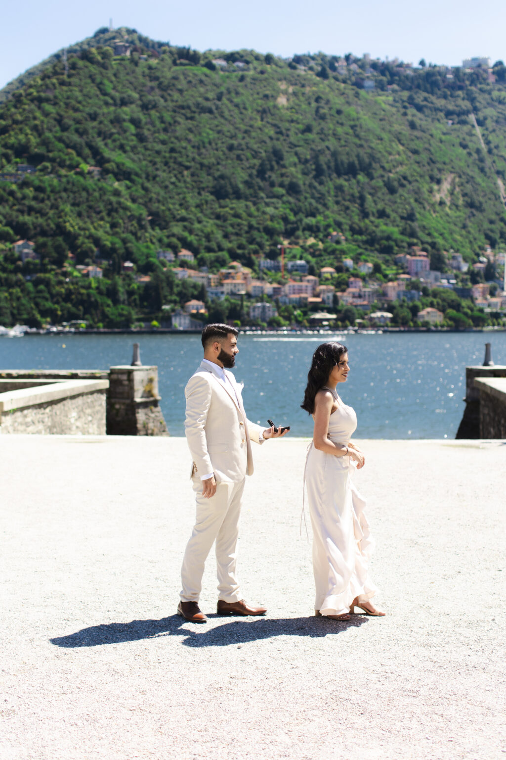 Man holding a ring box as his partner turns toward him on a lakefront terrace on Lake Como, the town of Como and Monte Brunate behind them