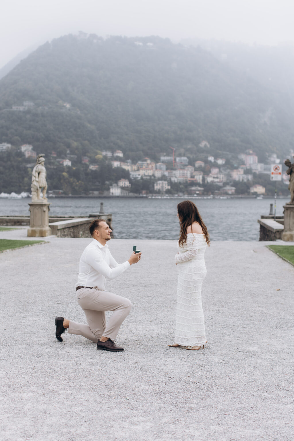 He kneels on one knee holding up the ring box to his pregnant partner, classical statues and the misty Como hillside behind them, in the gardens of Villa Olmo on Lake Como.