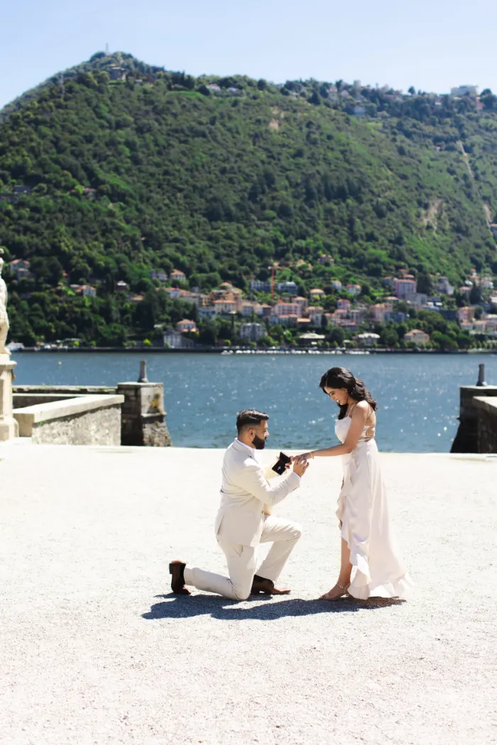 A man kneeling on one knee to propose on a lakeside terrace, with the town of Como and Monte Brunate rising across the water.