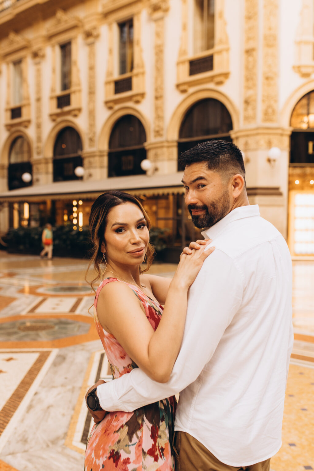 Mother in a floral dress and father in a white shirt share a close embrace inside the Galleria Vittorio Emanuele II during a quinceañera celebration in Milan.