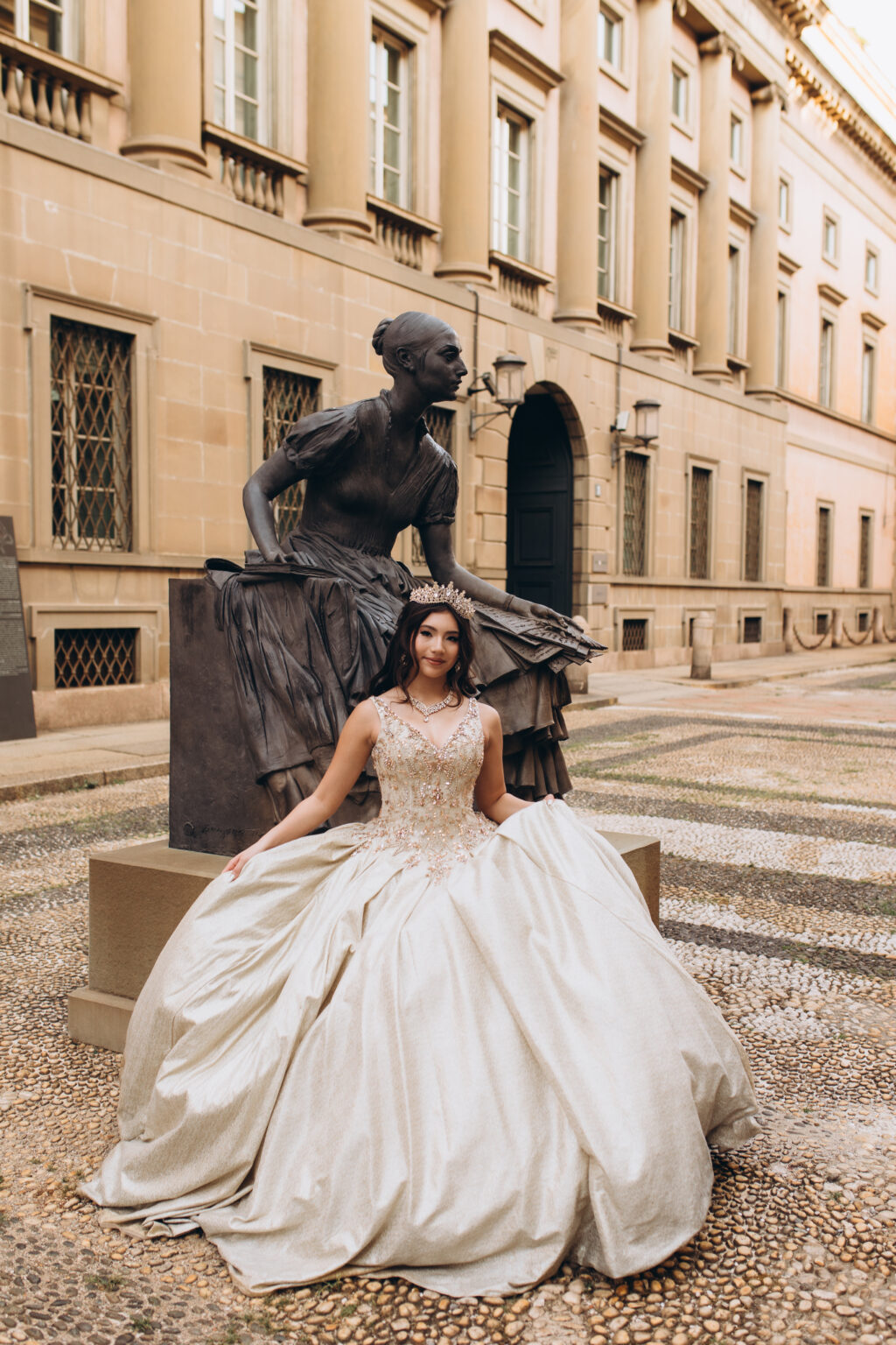 Quinceañera in a champagne gown sits beside a bronze statue of a seated woman before a historic palazzo on a cobbled Milan street.
