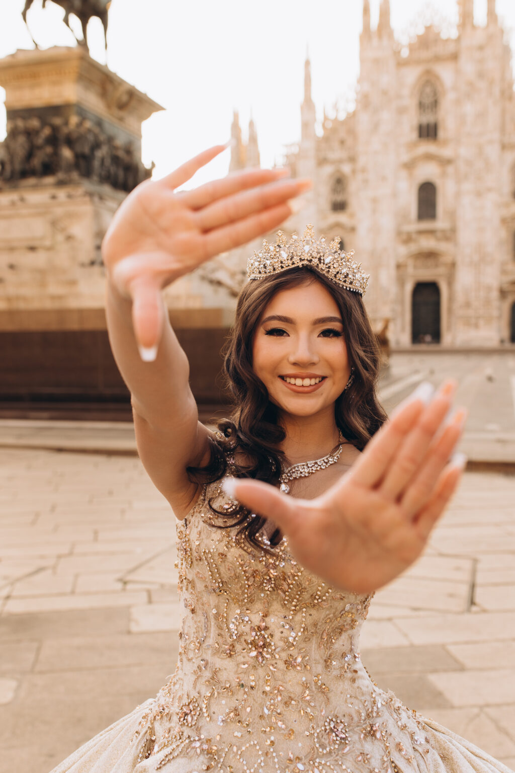 Crowned quinceañera framing her face with her hands near the equestrian monument to Victor Emmanuel II in Piazza del Duomo.