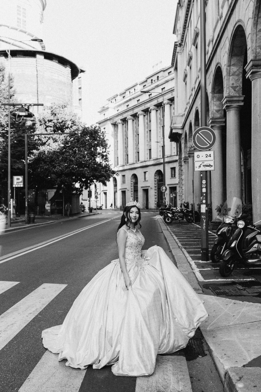 Black and white photo of a quinceañera seated on a crosswalk with her gown spread across a Milan street.