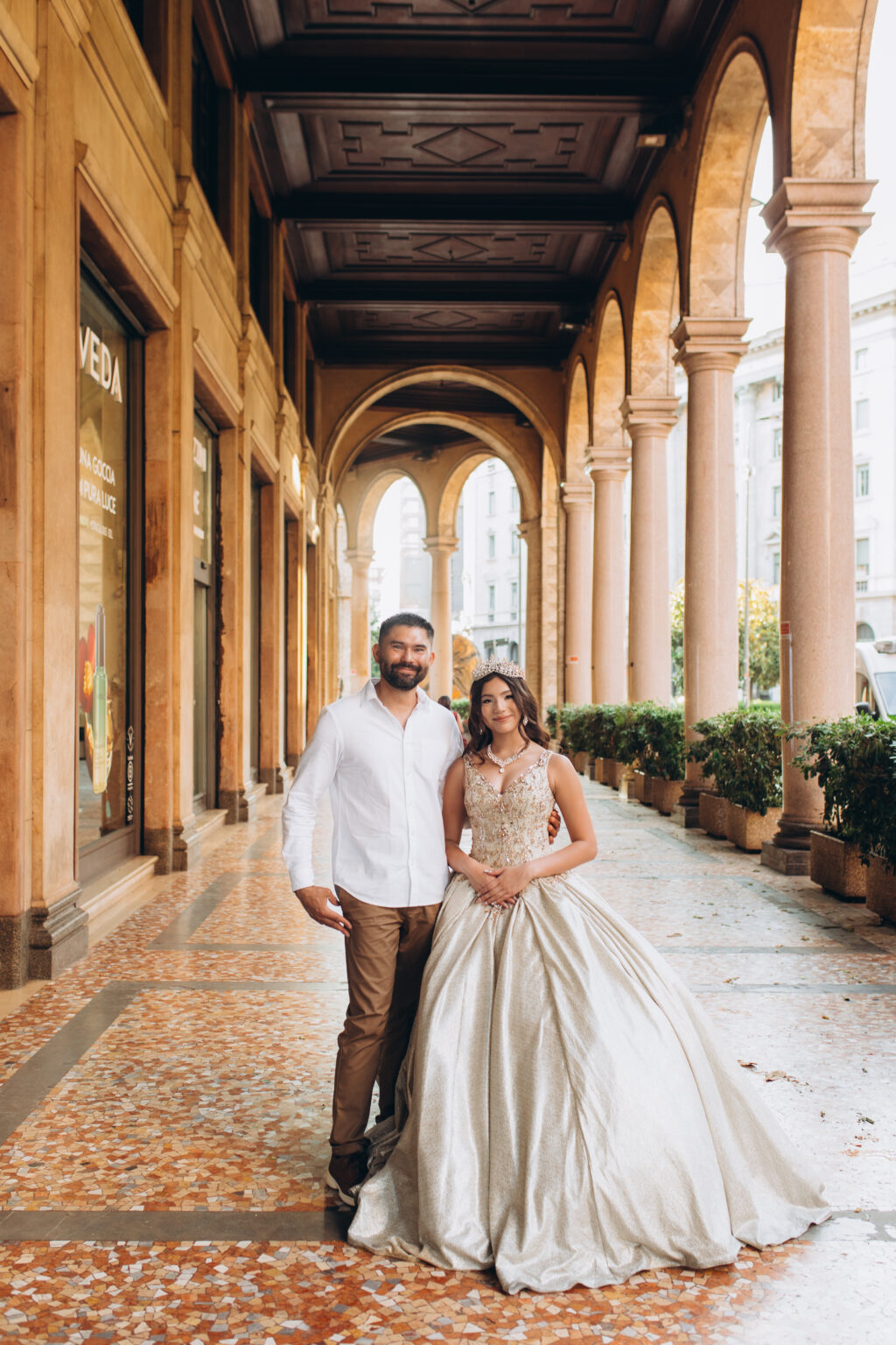 Quinceañera and her father stand together beneath the arched portico in central Milan during her quinceañera photoshoot in Milan.