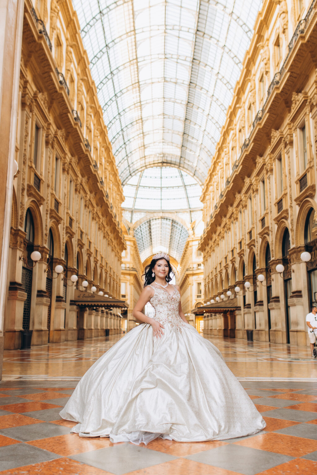 Quinceañera in a crown and champagne ball gown poses alone under the glass vault of the Galleria Vittorio Emanuele II in Milan.
