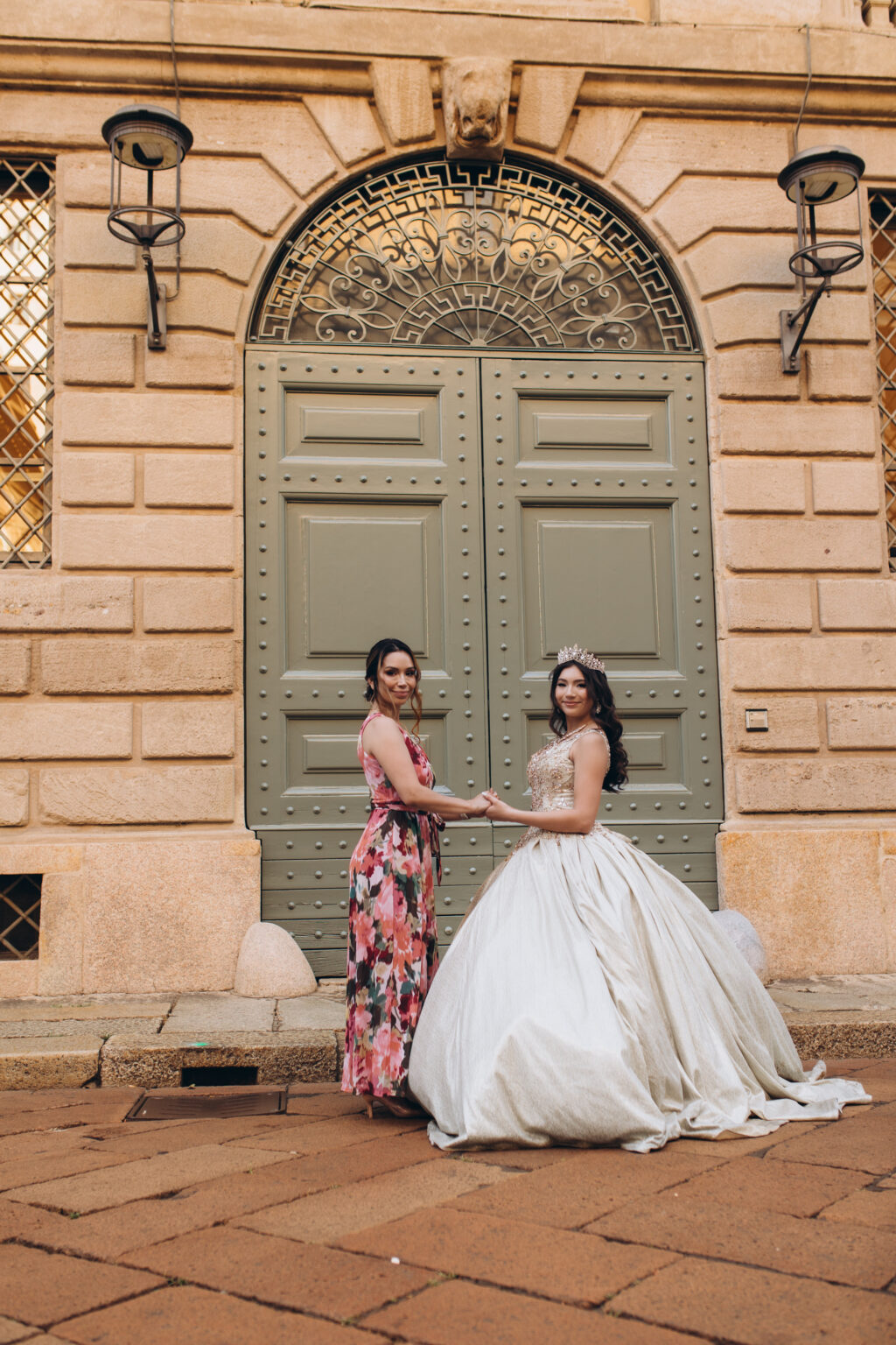 Quinceañera and her mother clasp hands before an ornate green palazzo door during a quinceañera photoshoot in Milan.