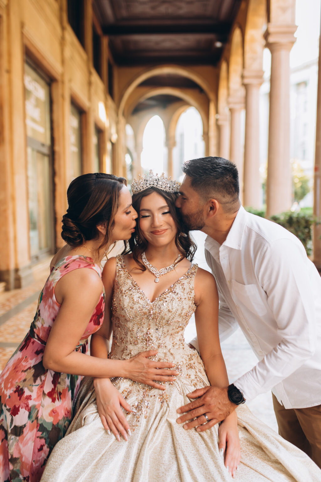 Mother and father each kiss their crowned quinceañera daughter on the cheek beneath a portico in central Milan.