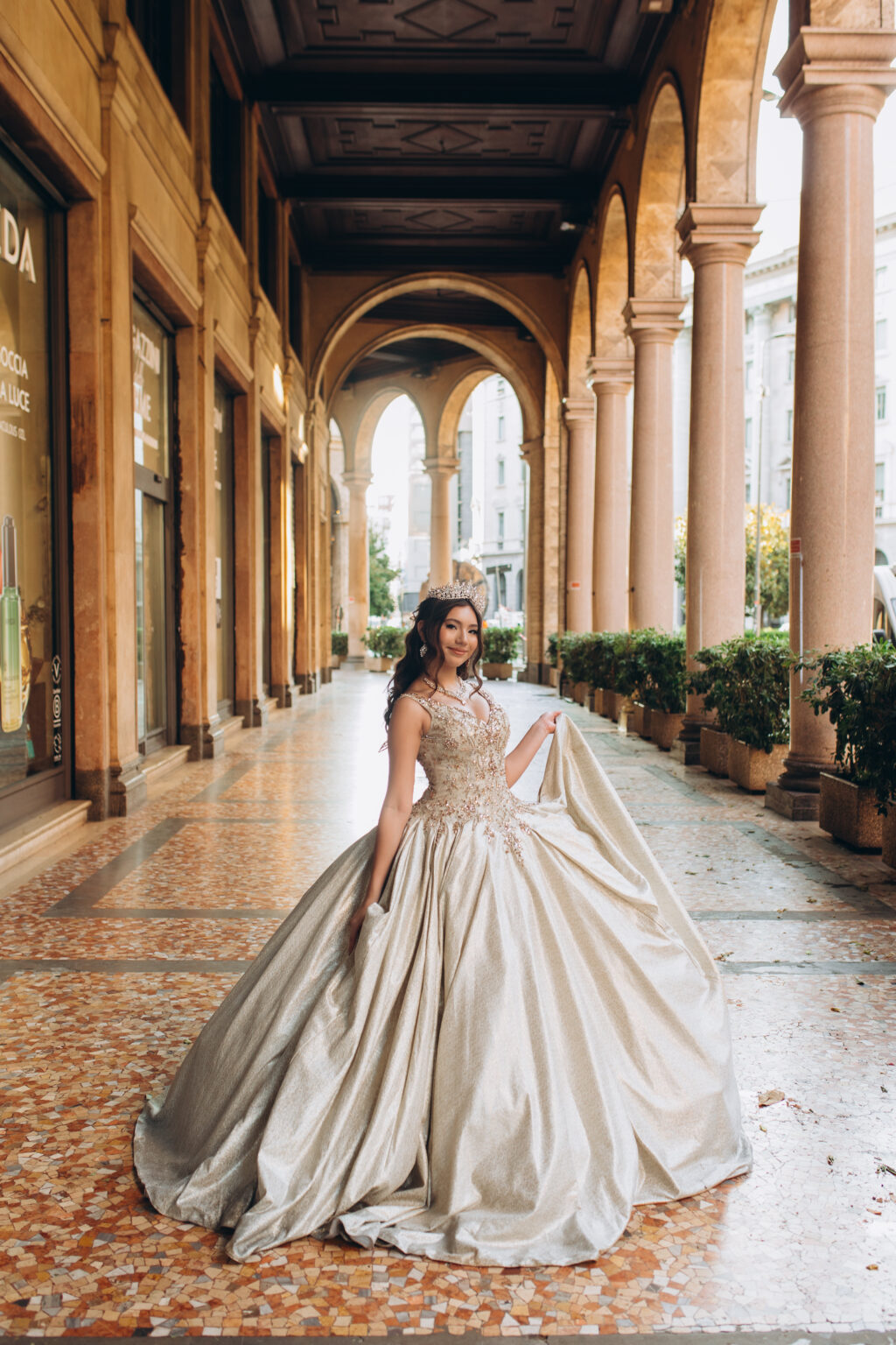 Quinceañera lifting her golden gown into a turn beneath the arches of a central Milan portico.