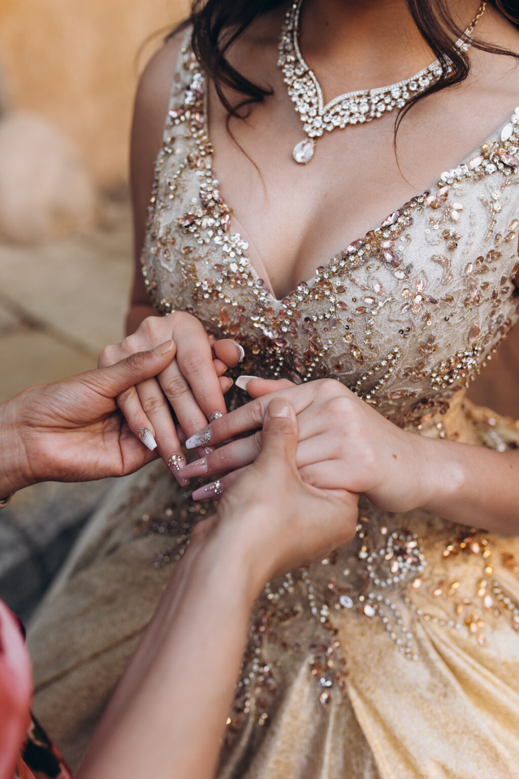 Detail of a mother's hands holding her quinceañera daughter's hands over a crystal-beaded bodice and diamond necklace in Milan.