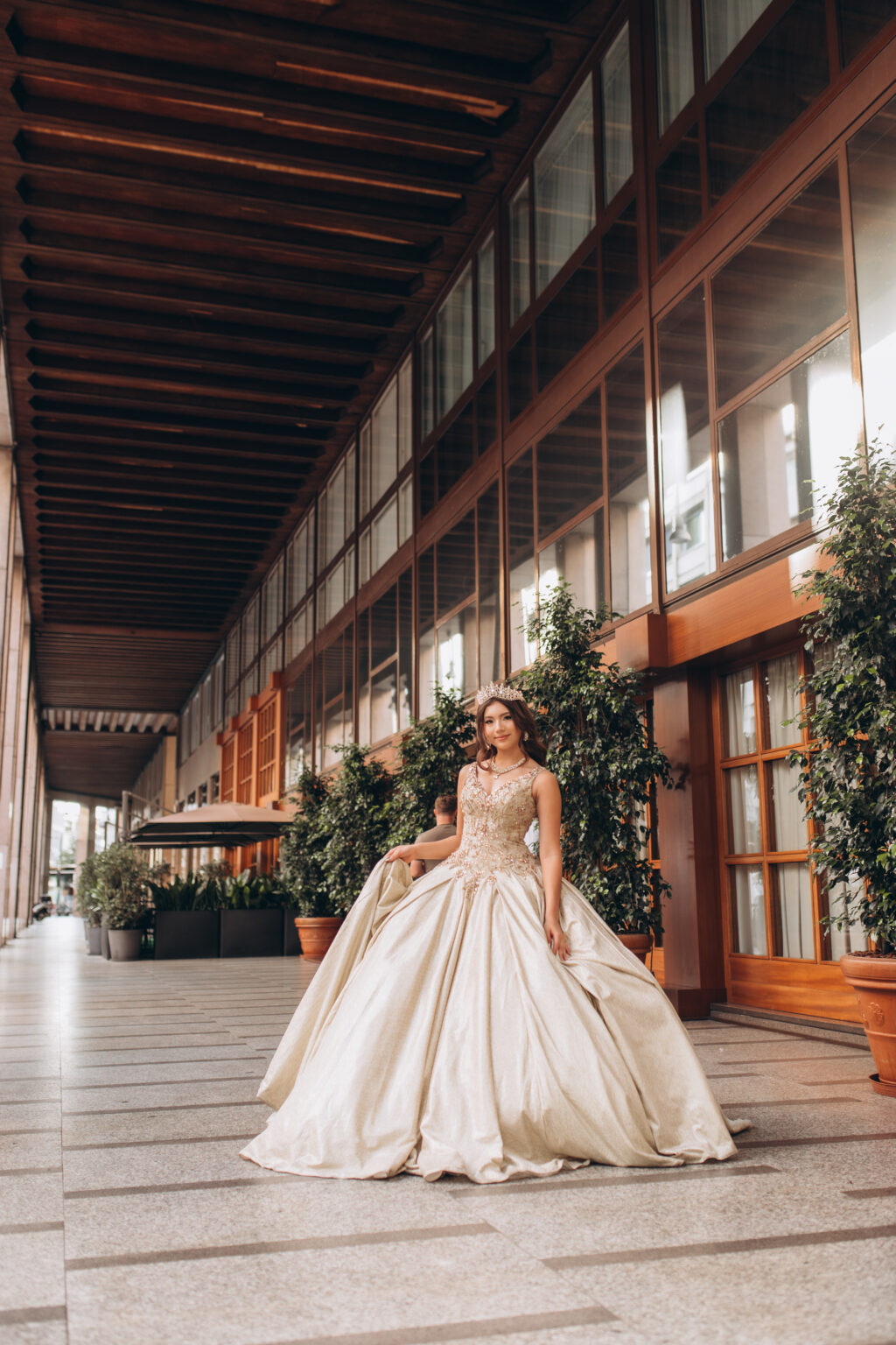 Quinceañera walking with her gown gathered beneath a modern wood-and-glass colonnade in Milan.