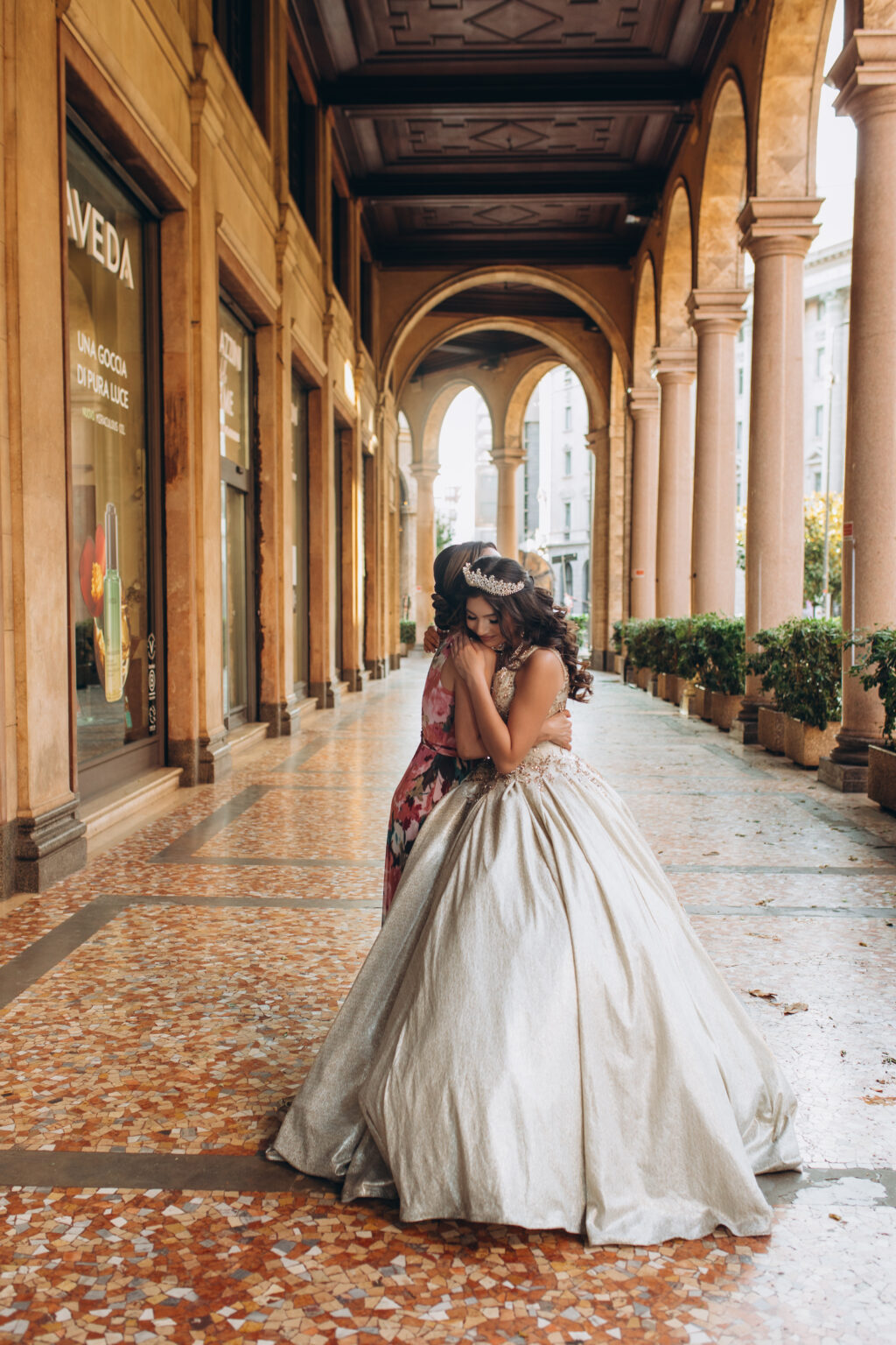 Quinceañera in a golden gown embraces her mother beneath an arched portico in central Milan during her quinceañera photoshoot in Milan.