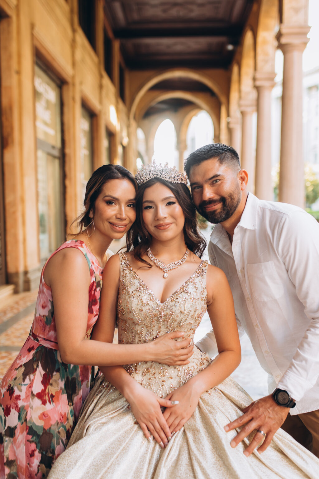 Quinceañera seated between her smiling mother and father under an arched portico in central Milan.
