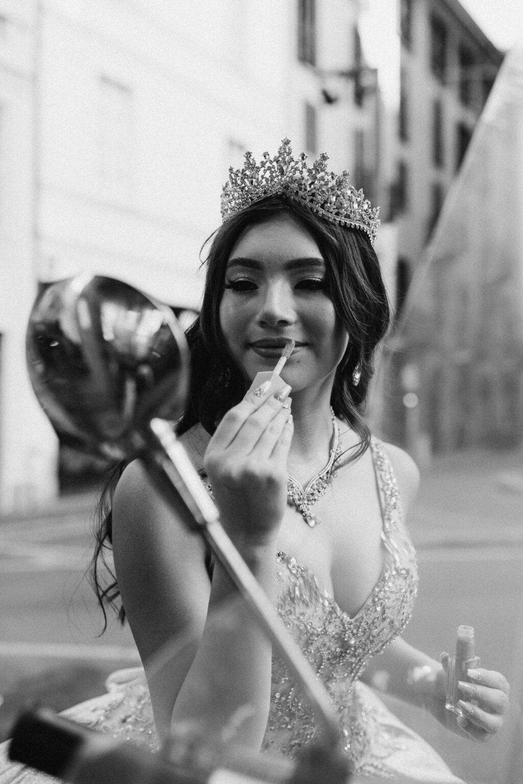 Black and white candid of a crowned quinceañera applying lip gloss on a Milan street. Заголовок: Last Touches
