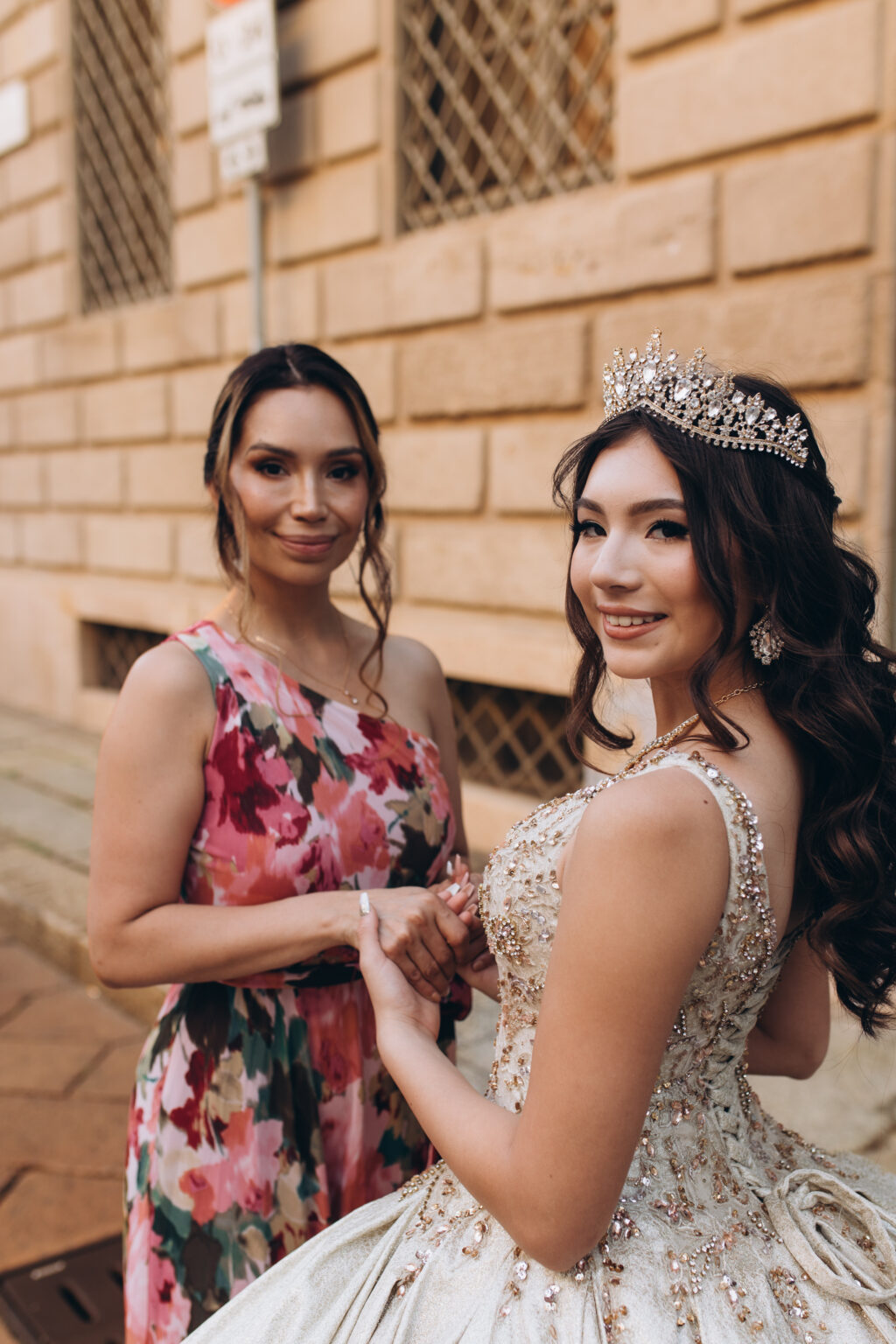 Close portrait of a quinceañera and her mother holding hands against a rusticated stone palazzo wall in Milan.