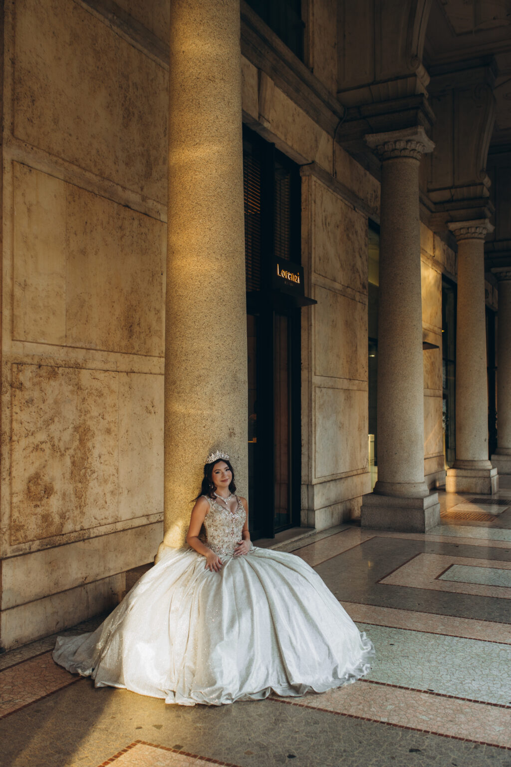 Quinceañera resting against a tall column with her gown pooled across the terrazzo floor of a central Milan portico.