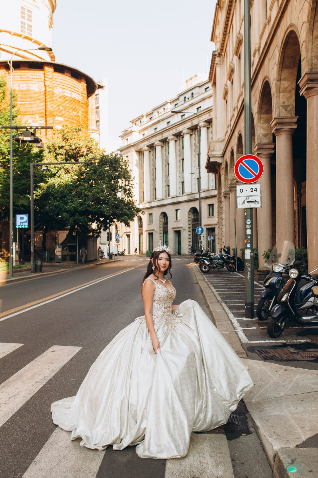 Quinceañera in a crown stands on a crosswalk on a quiet Milan street lined with scooters, a historic rotunda rising behind her.