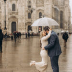 A couple kisses under a clear umbrella on rain-soaked Piazza del Duomo with the cathedral behind them in Milan