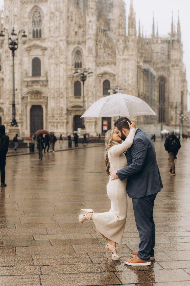 A couple kisses under a clear umbrella on rain-soaked Piazza del Duomo with the cathedral behind them in Milan