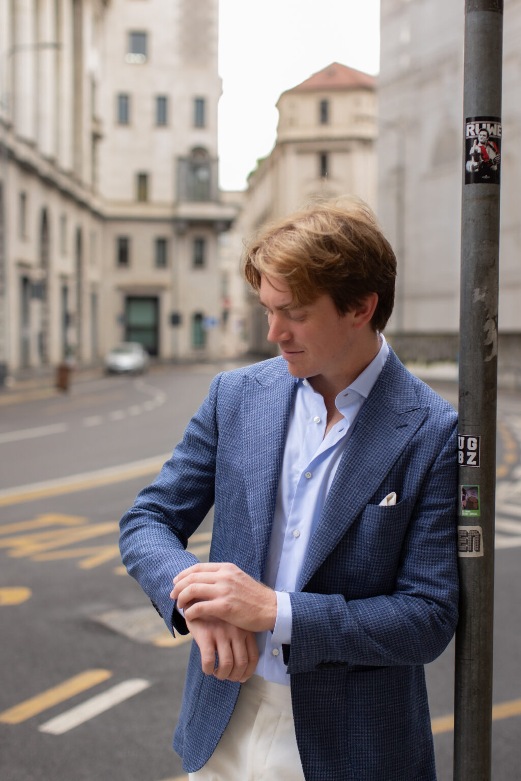 A man in a blue jacket leaning against a lamp post on a quiet Milan street, glancing down at his watch.