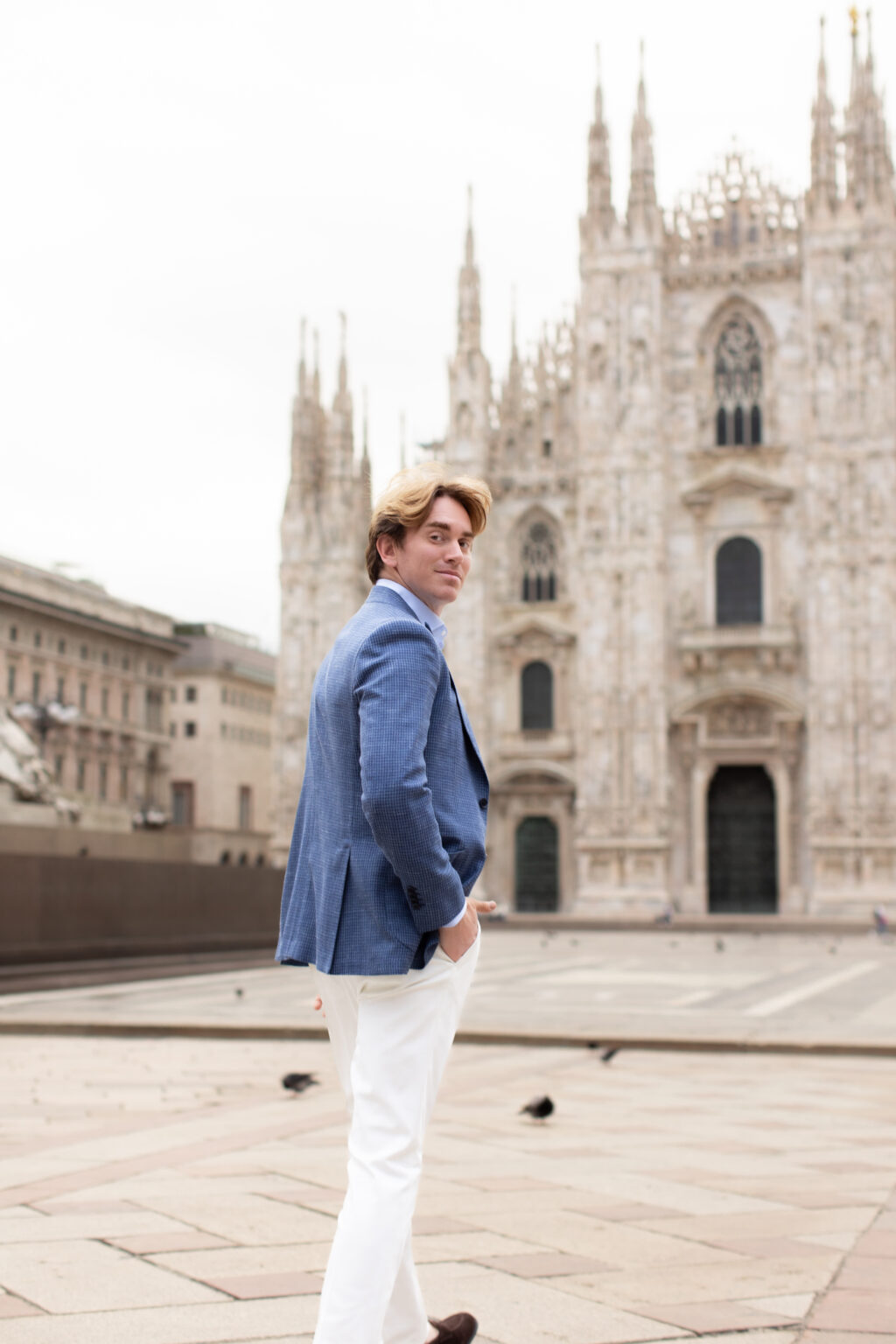 A man in a blue blazer glancing back over his shoulder while walking away, with the Duomo di Milano behind him in Milan.
