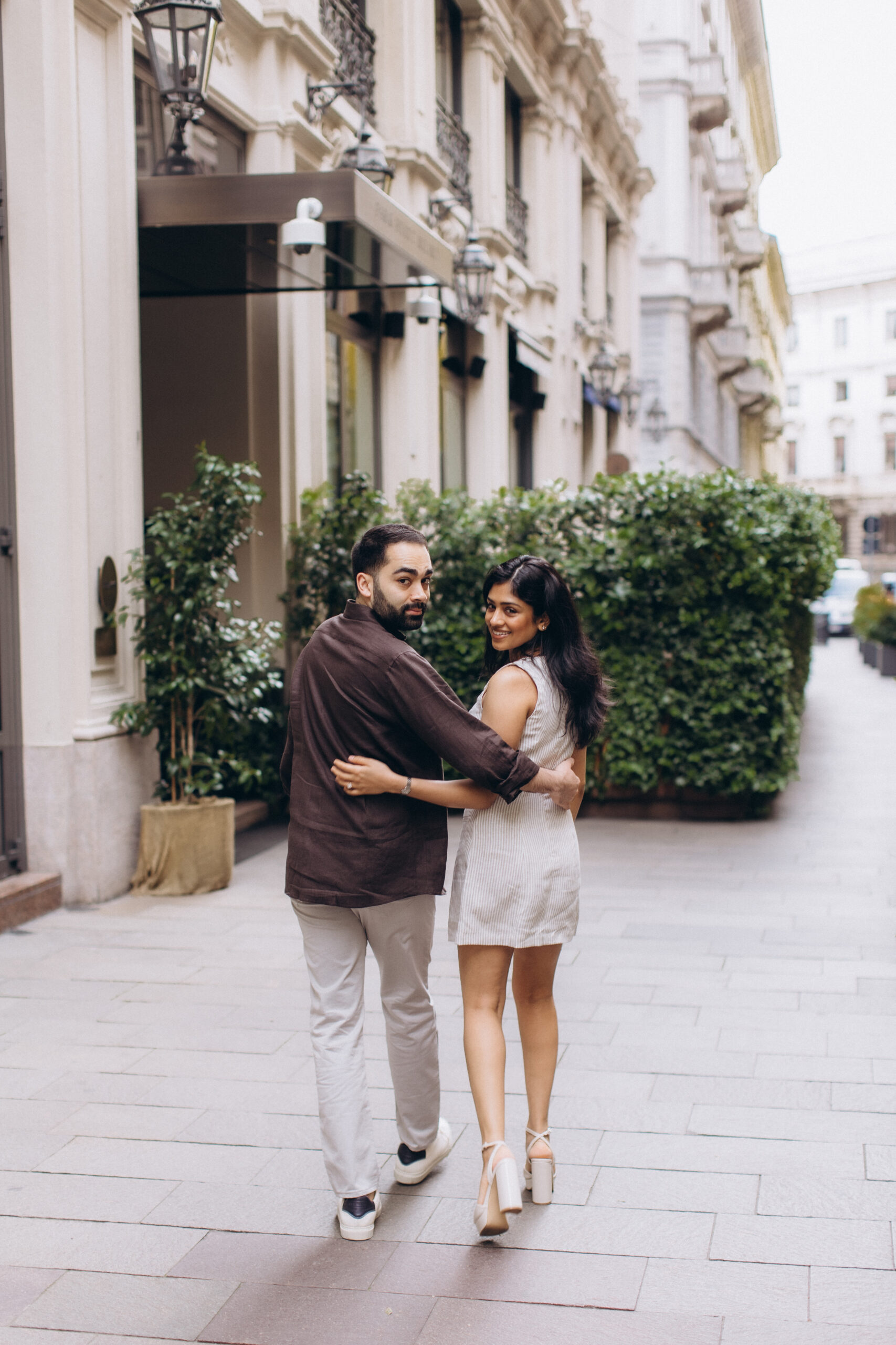 Summer Couple Photoshoot on a Milan Street A couple walks arm in arm down a Milan street lined with green hedges and looks back over their shoulders on a summer day