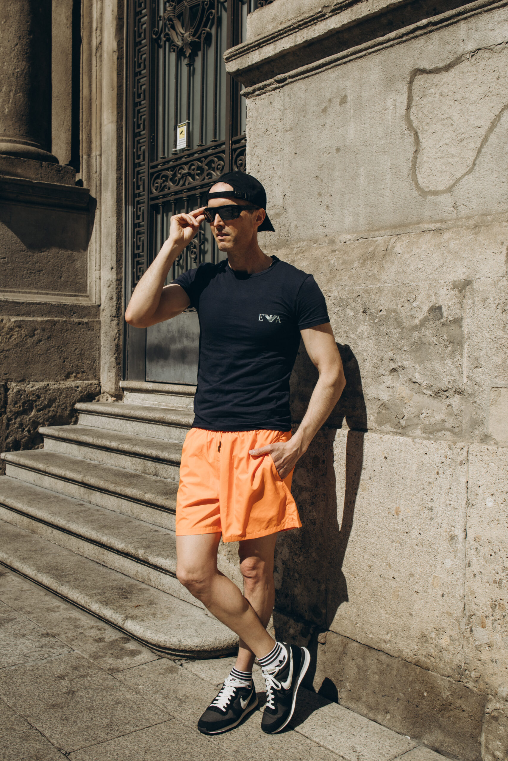 Summer Midday Portrait in Milan A man in a black tee and orange shorts leans against a stone wall by an ornate iron door in bright midday sun in Milan