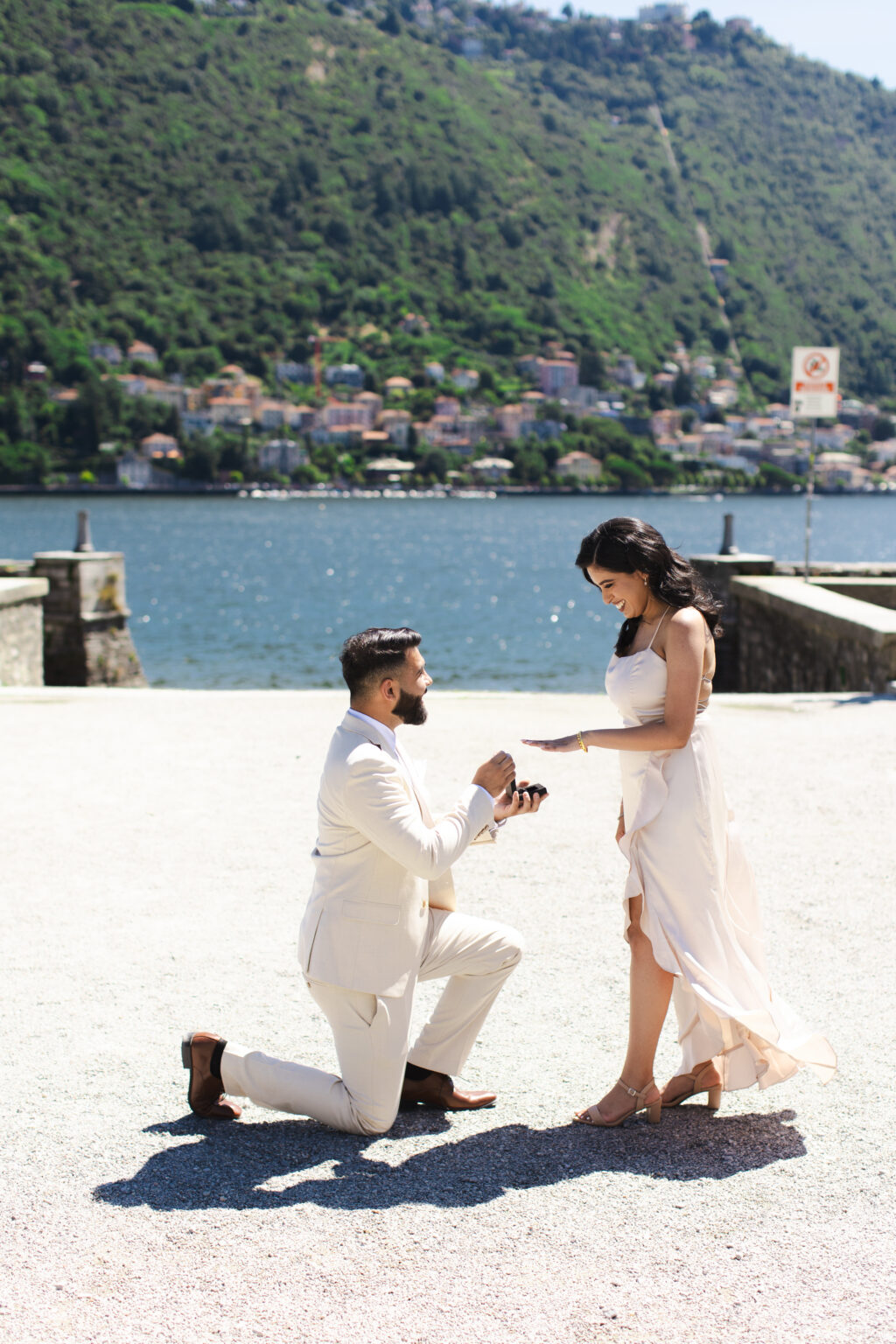 Man on one knee proposing with a ring as his astonished partner reacts on a lakefront terrace on Lake Como, the lake and hills behind them
