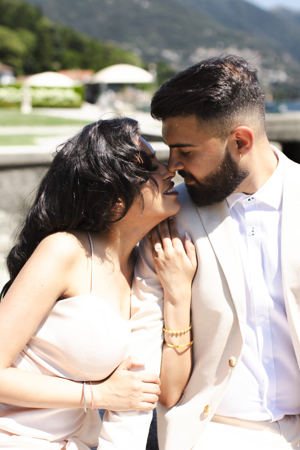 A tender close-up of a newly engaged couple about to kiss, the diamond ring on her hand resting on his chest, in a lakeside villa garden on Lake Como.