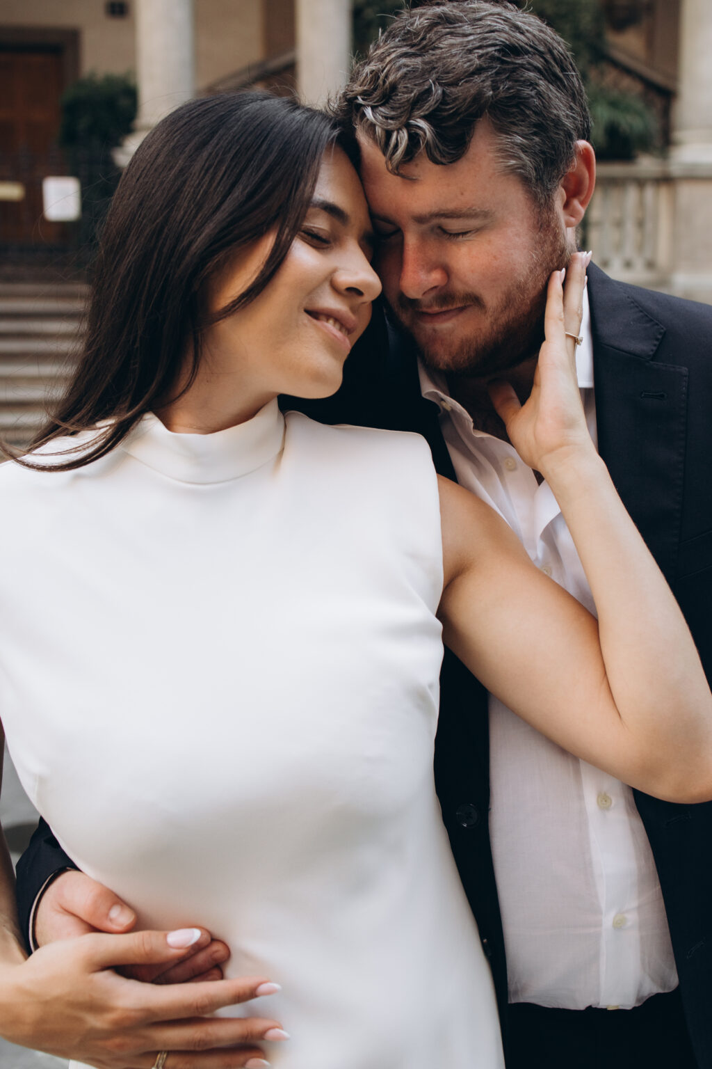 Newly engaged couple embracing forehead to forehead with eyes closed, the woman's hand resting on her fiance's face, in a Milan courtyard